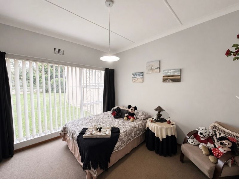 A well-maintained bedroom featuring neutral carpeting, a large window with vertical blinds, and a central pendant light fixture.