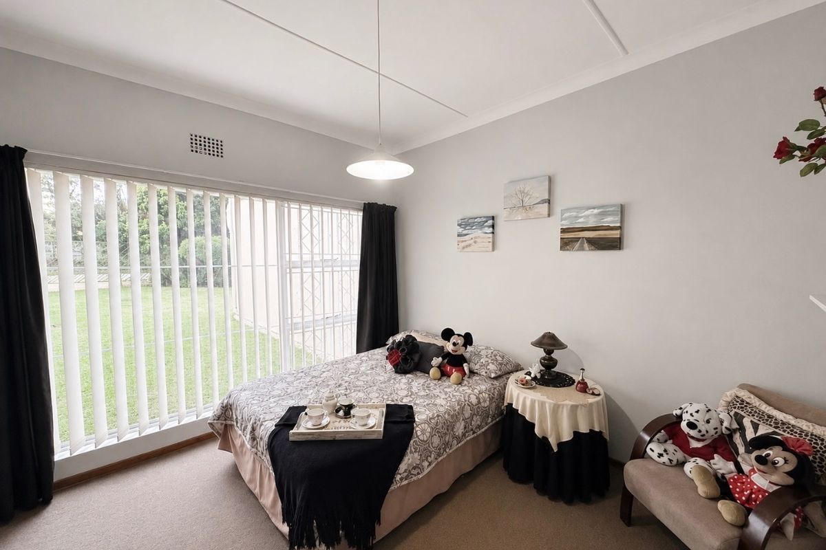 A well-maintained bedroom featuring neutral carpeting, a large window with vertical blinds, and a central pendant light fixture.