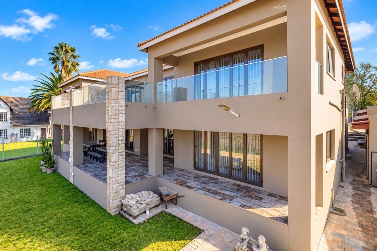 A spacious two-level rear exterior featuring a covered tiled patio, glass balustrades, and stone-clad architectural detailing overlooking a landscaped garden.