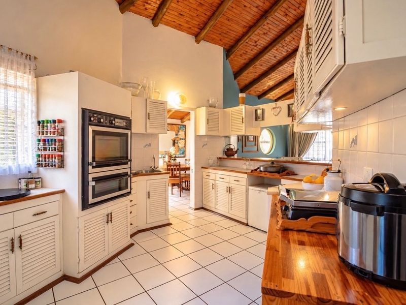 A rustic kitchen featuring exposed timber beam ceilings, white louvered cabinetry, timber countertops, and a built-in double oven with tiled flooring.