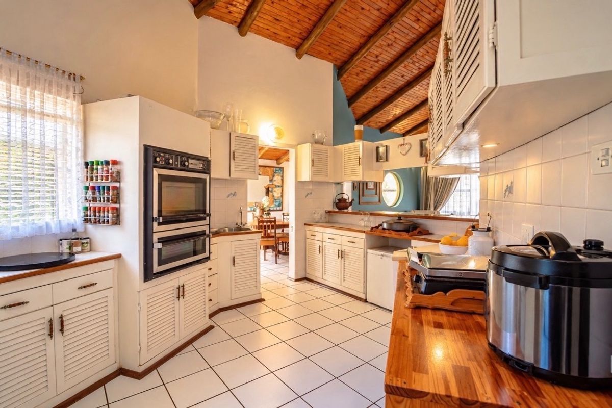 A rustic kitchen featuring exposed timber beam ceilings, white louvered cabinetry, timber countertops, and a built-in double oven with tiled flooring.