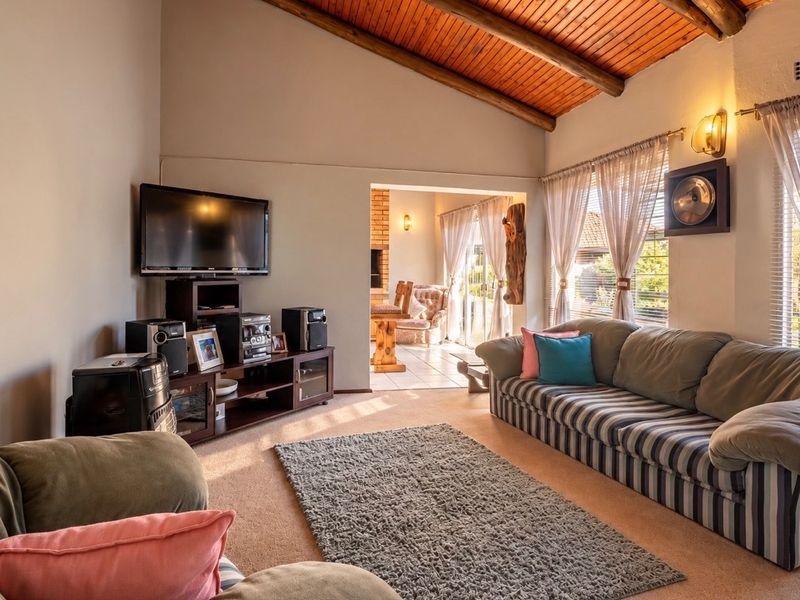 A cozy living room featuring a vaulted ceiling with exposed timber beams, carpeted flooring, and natural light flowing through the windows.