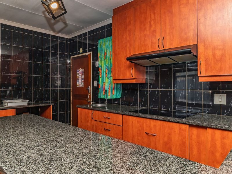 A functional kitchen featuring granite countertops, wooden cabinetry, an electric hob with extractor fan, and dark tiled wall finishes.