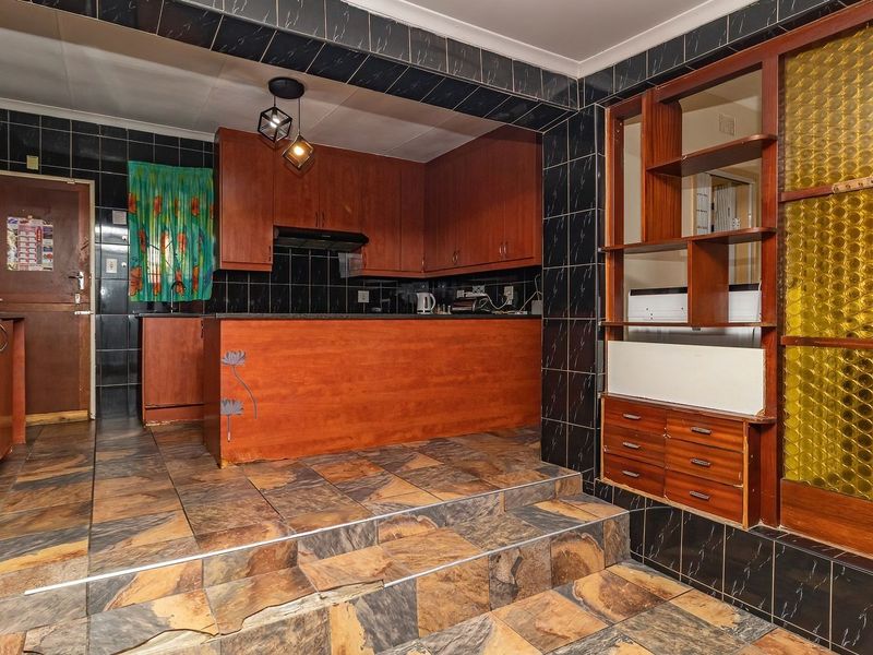 A kitchen featuring wooden cabinetry, black tiled walls, and a raised floor area with stone-look tiles, including a built-in oven and integrated shelving.