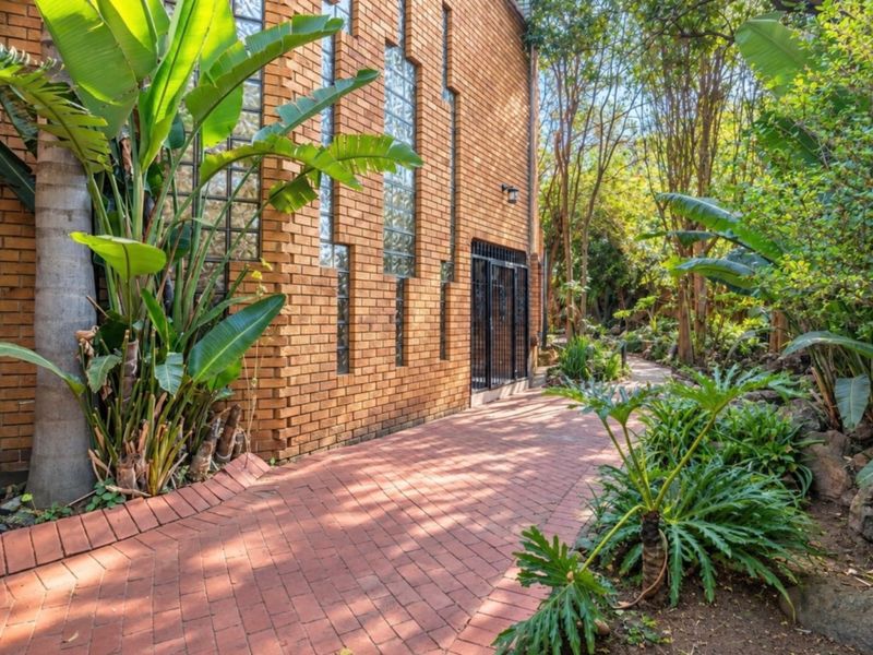 A well-maintained rear garden path featuring brick paving, face brick exterior walls with glass block accents, and lush established landscaping.
