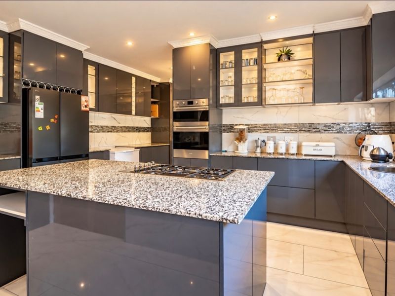 Modern kitchen featuring high-gloss dark cabinetry, granite countertops, a central island with a gas hob, built-in appliances, and elegant glass-fronted display cabinets.