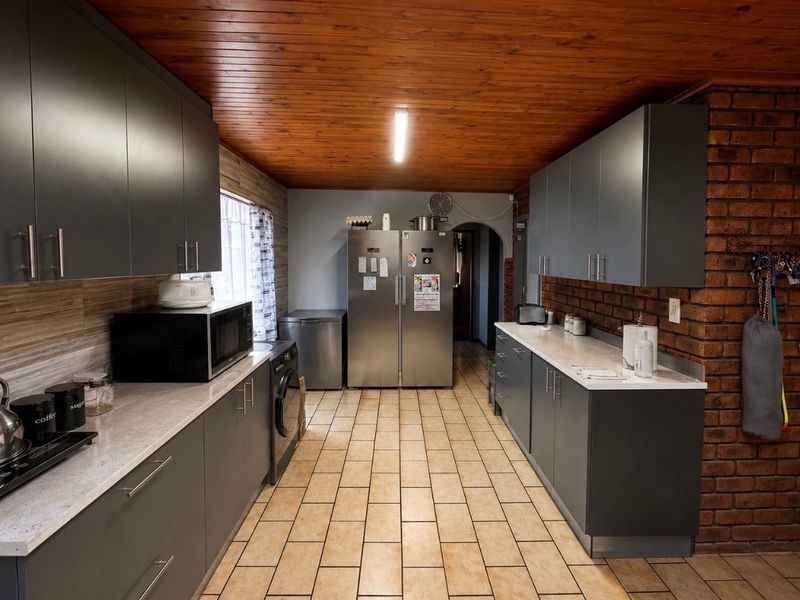 A functional kitchen featuring modern grey cabinetry, stone-look countertops, exposed brick accents, and a warm timber ceiling with tiled flooring.