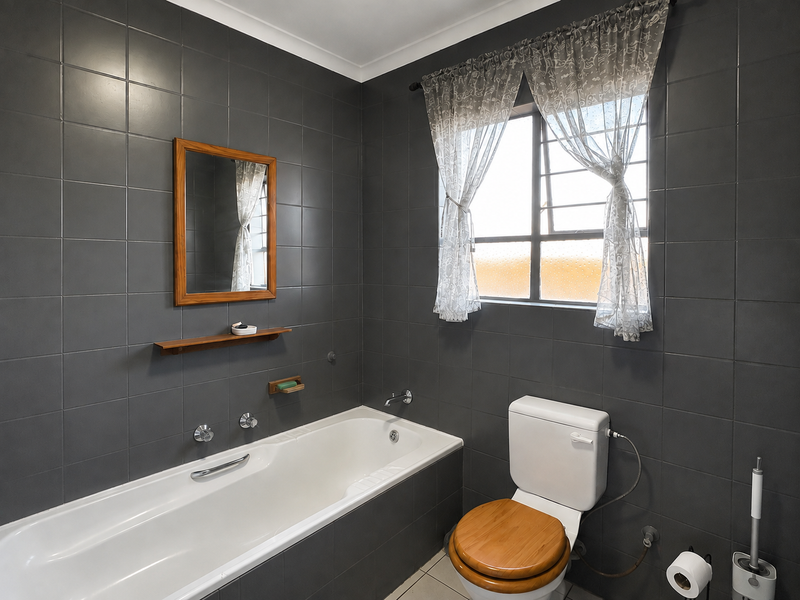 A modern bathroom featuring dark grey wall tiles, a white built-in bathtub, and a toilet, complemented by a window with security bars.