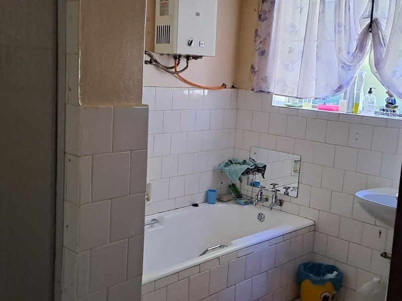 A dated bathroom featuring a standard bathtub, pedestal basin, and wall-mounted gas geyser with visible tile finishes.