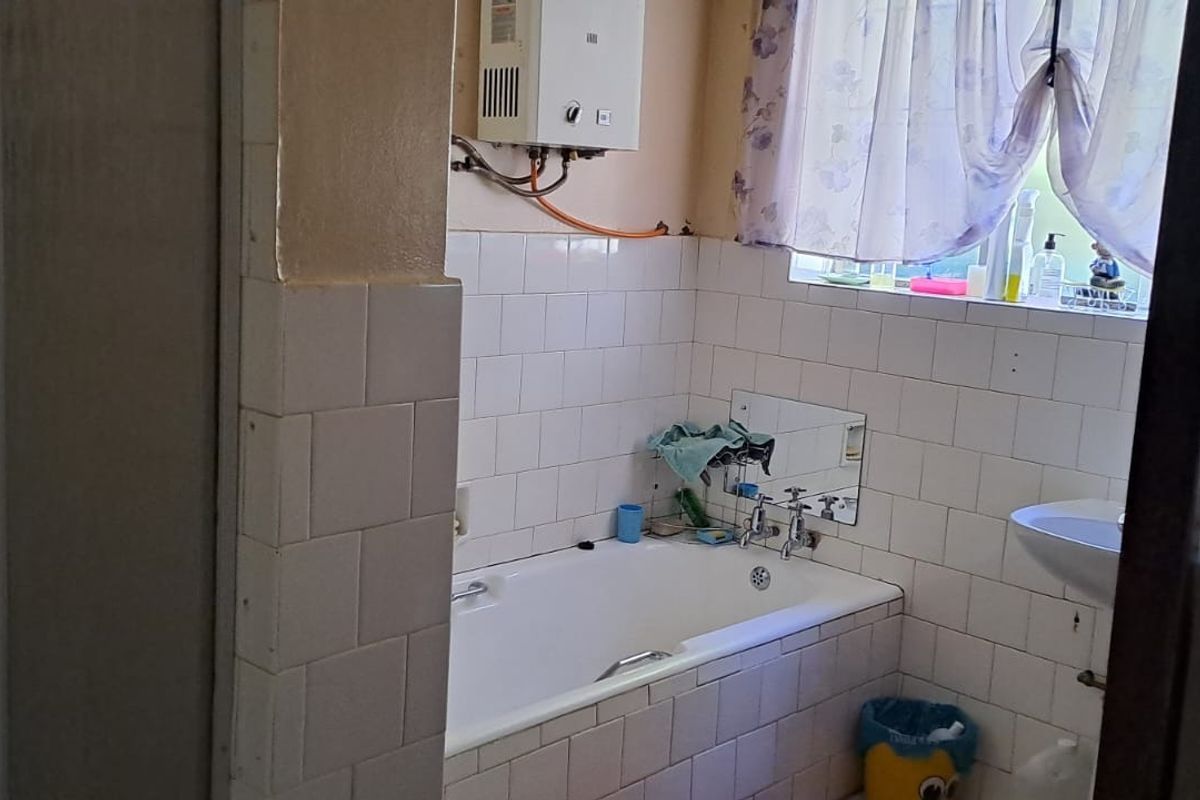 A dated bathroom featuring a standard bathtub, pedestal basin, and wall-mounted gas geyser with visible tile finishes.