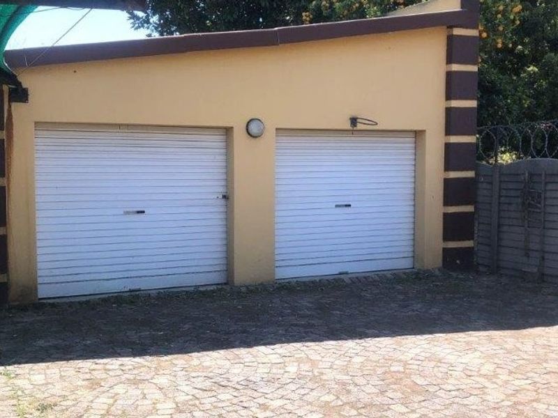 A double garage featuring white roller doors, set within a paved driveway area enclosed by precast walling and security fencing.