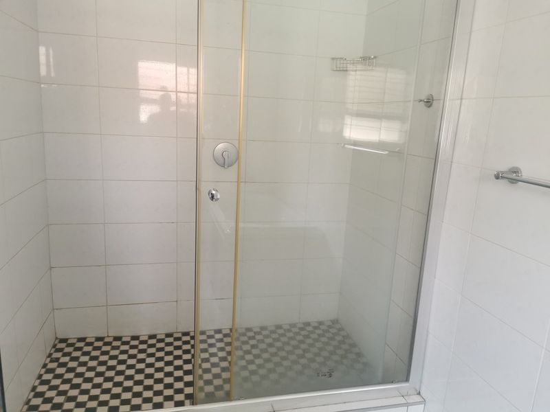 A clean bathroom shower enclosure featuring white wall tiles, a checkered floor pattern, and a glass door with gold-tone trim.