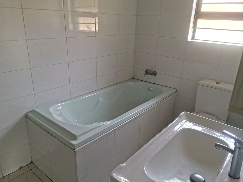 A functional bathroom featuring a built-in bathtub, pedestal basin, and toilet, finished with white ceramic wall and floor tiles.