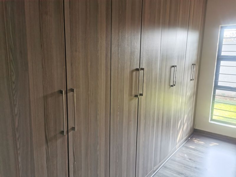 A bright bedroom featuring modern wood-finish built-in cupboards, laminate flooring, and a large window providing natural light.