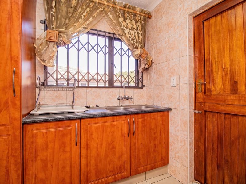 A functional scullery area featuring built-in wooden cabinetry, a double sink, tiled wall finishes, and a secure window with metal burglar bars.