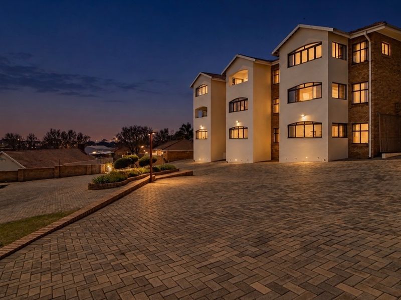 A well-maintained apartment complex exterior at dusk, featuring paved driveway access, multi-story brick and plaster facade, and integrated exterior lighting.