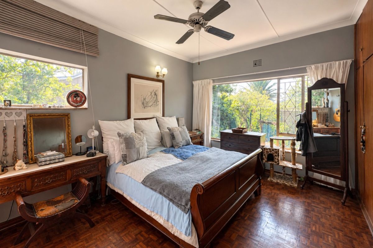 A well-maintained bedroom featuring classic parquet flooring, a ceiling fan, and large windows providing natural light and garden views.