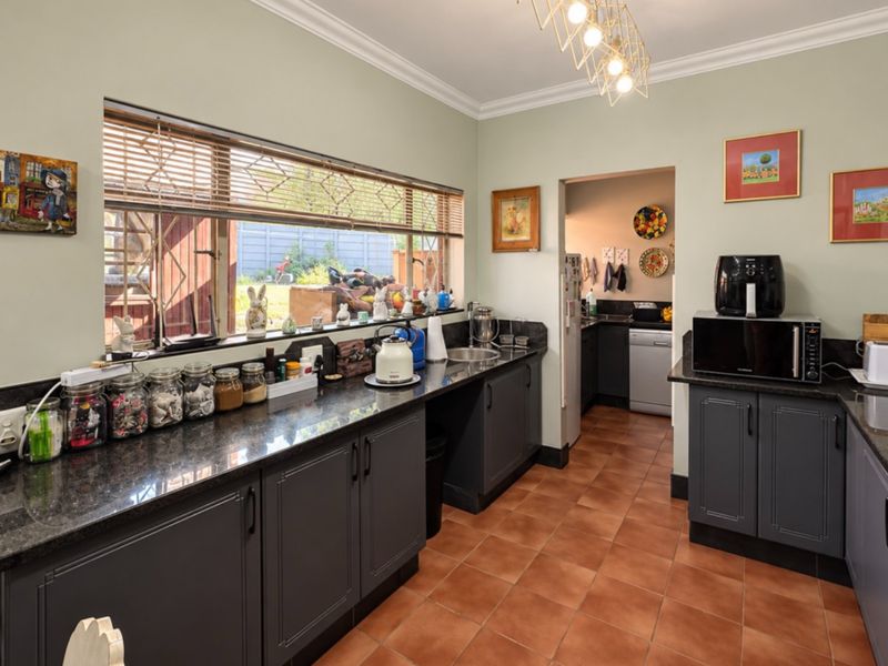 A functional kitchen featuring dark grey cabinetry, granite countertops, terracotta flooring, and a convenient adjoining scullery area with natural light.
