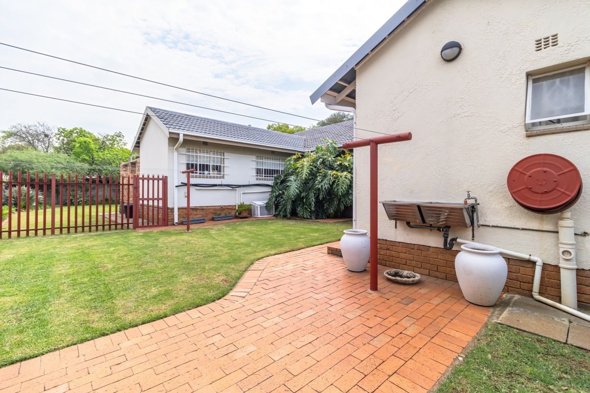 Rear exterior view featuring a brick-paved patio area, lawn, metal security fencing, and a wall-mounted utility wash trough.