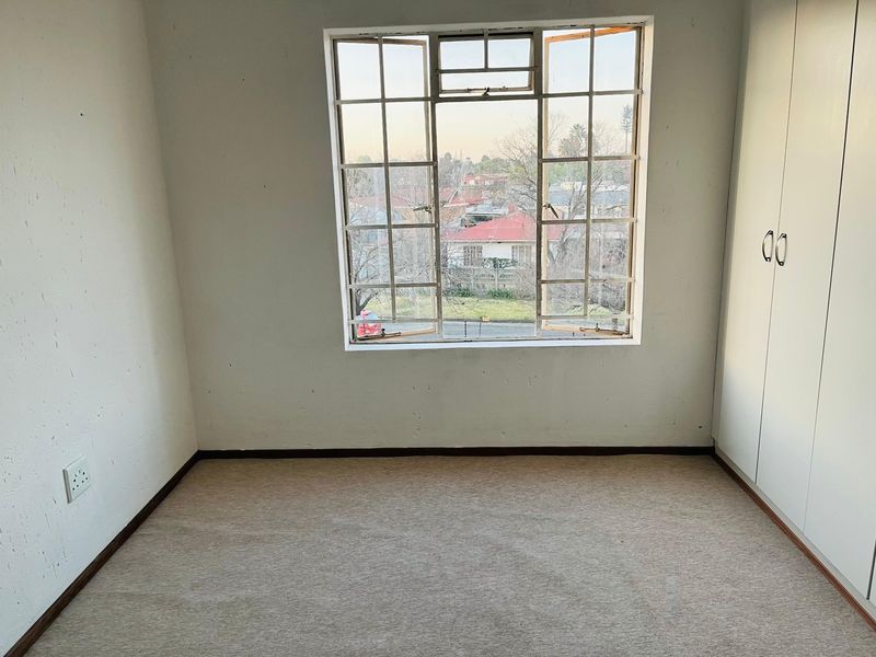 An empty bedroom featuring built-in cupboards, carpeted flooring, and a large steel-framed window with views of the surrounding neighborhood.