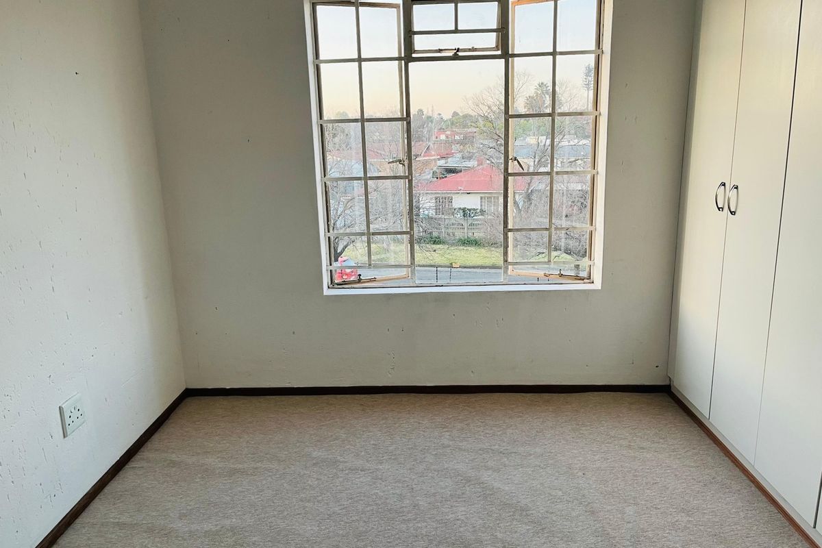An empty bedroom featuring built-in cupboards, carpeted flooring, and a large steel-framed window with views of the surrounding neighborhood.