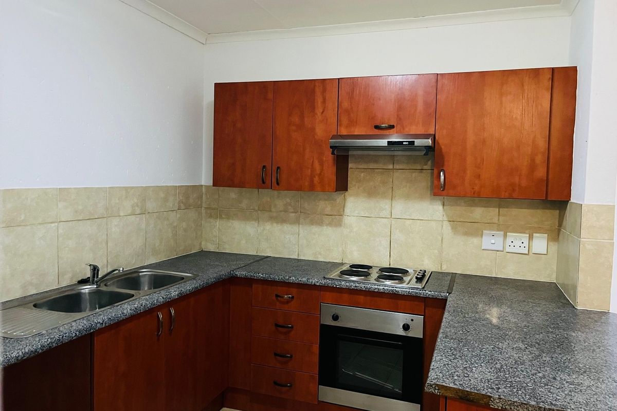 A functional kitchen featuring wood-finish cabinetry, granite-look countertops, a built-in oven, electric hob, and a stainless steel double sink with tiled backsplash.