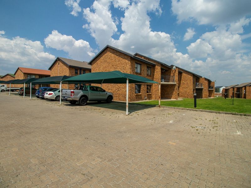 Exterior view of a brick apartment complex featuring paved parking bays with shade cloth carports and landscaped lawn areas.