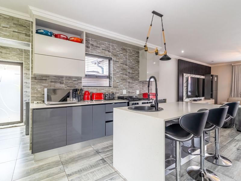 Modern open-plan kitchen featuring high-gloss cabinetry, stone-look tiled backsplash, a central island with a sink, and contemporary pendant lighting.