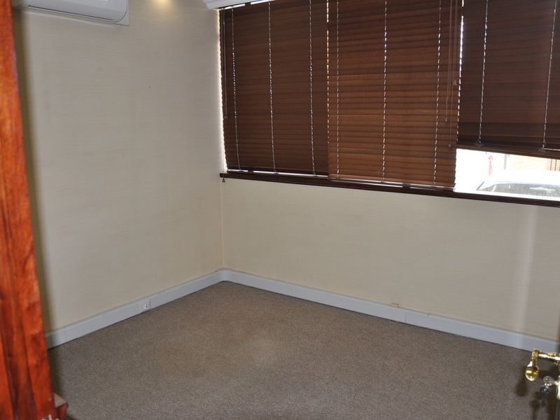 A vacant office space featuring carpeted flooring, a wall-mounted air conditioning unit, and dark wood-toned venetian blinds.