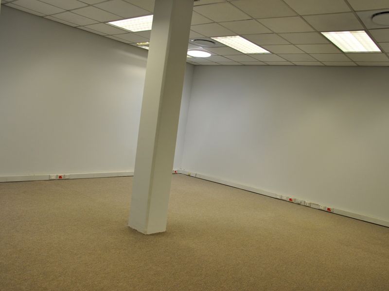 An empty, open-plan office space featuring a suspended ceiling, recessed lighting, carpeted flooring, and perimeter trunking for electrical and data connectivity.