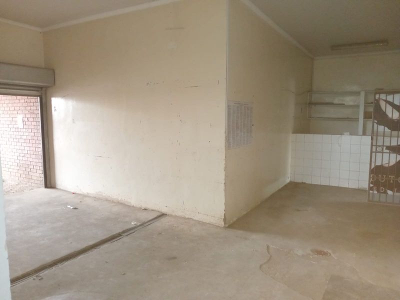 An empty open-plan retail space featuring concrete flooring, built-in shelving, a tiled splashback area, and a secure roller shutter entrance.