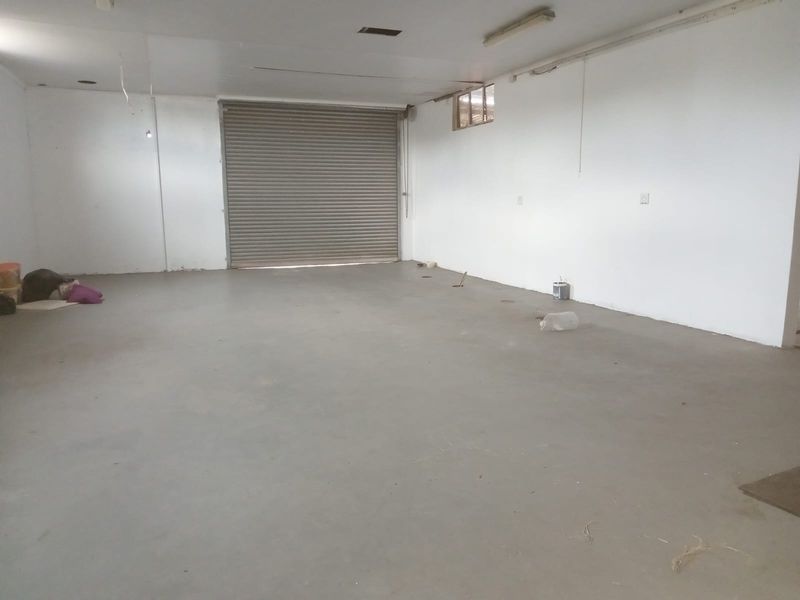 A spacious, vacant retail or garage area featuring a secure roller shutter door, concrete flooring, and basic utility lighting.
