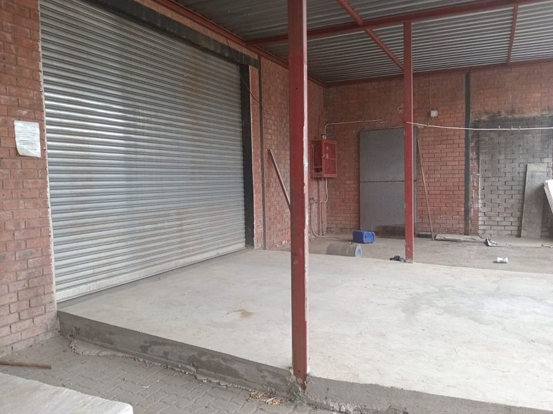 A functional retail exterior space featuring a secure roller shutter door, exposed brick walls, and a covered concrete loading or entrance area.