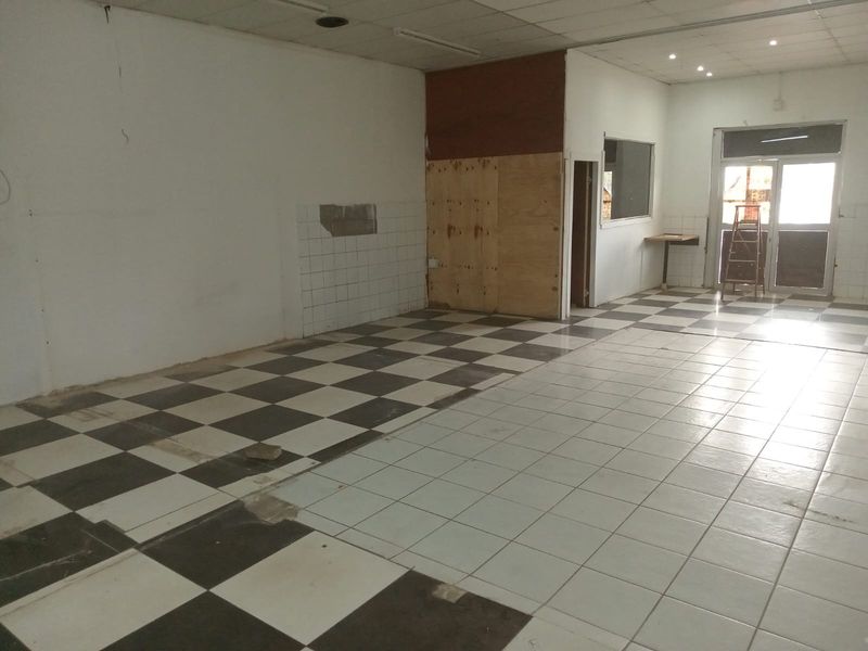 An empty, open-plan retail space featuring a checkered tiled floor, suspended ceiling, and glass entrance door, currently requiring renovation and cosmetic repairs.