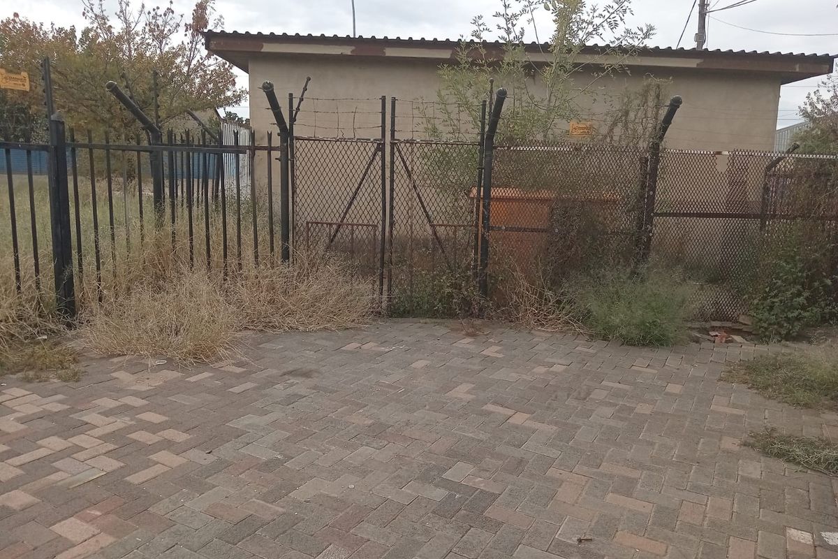 Rear exterior view of a retail property featuring a paved area, metal security fencing with barbed wire, and a structure with a corrugated metal roof.