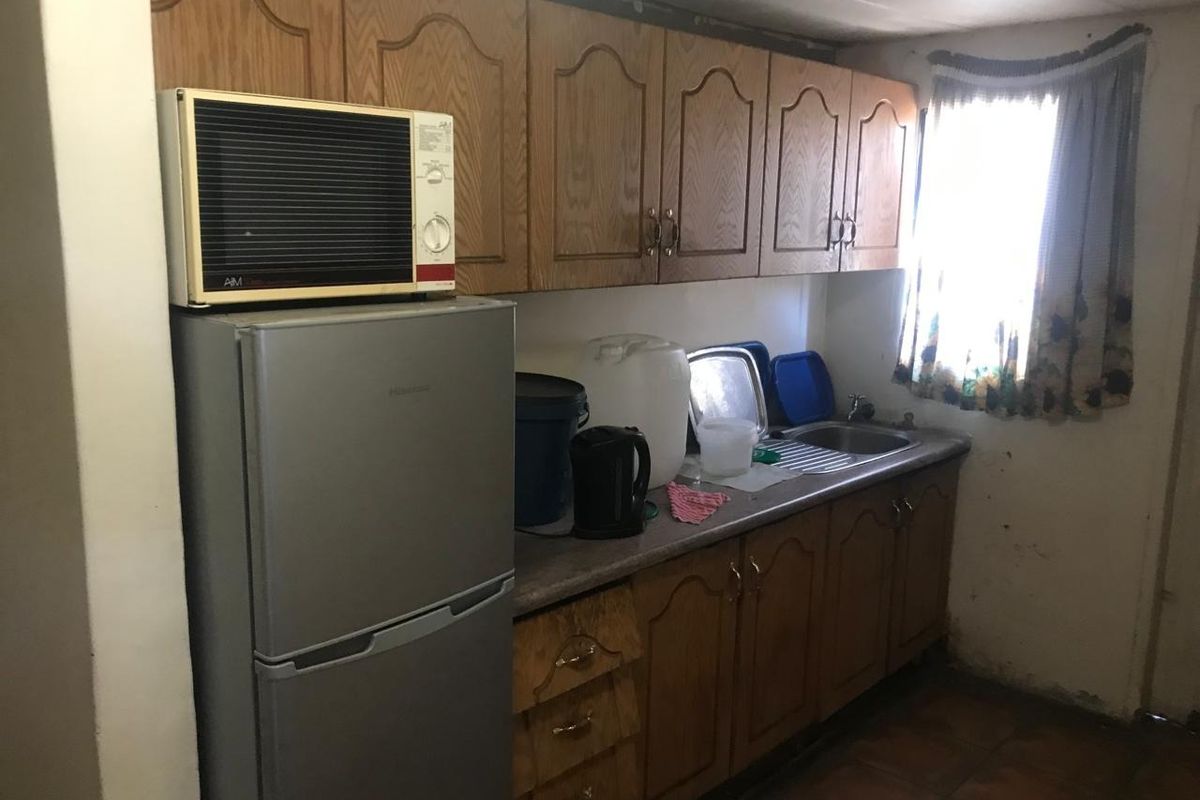 A compact kitchen featuring dated wooden cabinetry, a stainless steel sink, and tiled flooring, requiring cosmetic updates to improve its overall appeal.