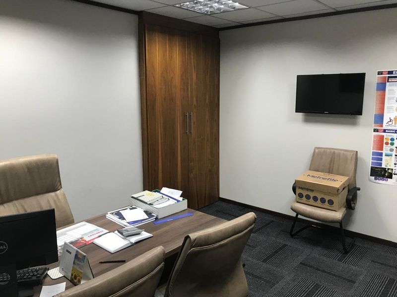 A functional office space featuring a built-in wooden storage cupboard, suspended ceiling with recessed lighting, and dark carpeted flooring.