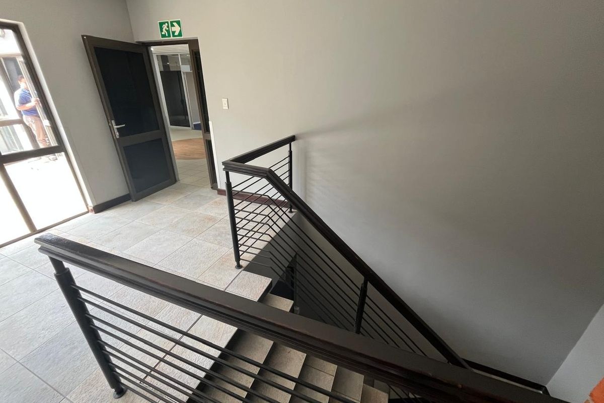 A clean, modern office hallway featuring tiled flooring and a metal staircase railing leading to the lower level.