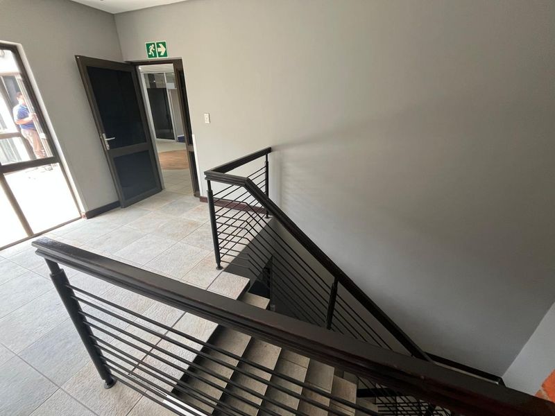 A clean, modern office hallway featuring tiled flooring and a metal staircase railing leading to the lower level.