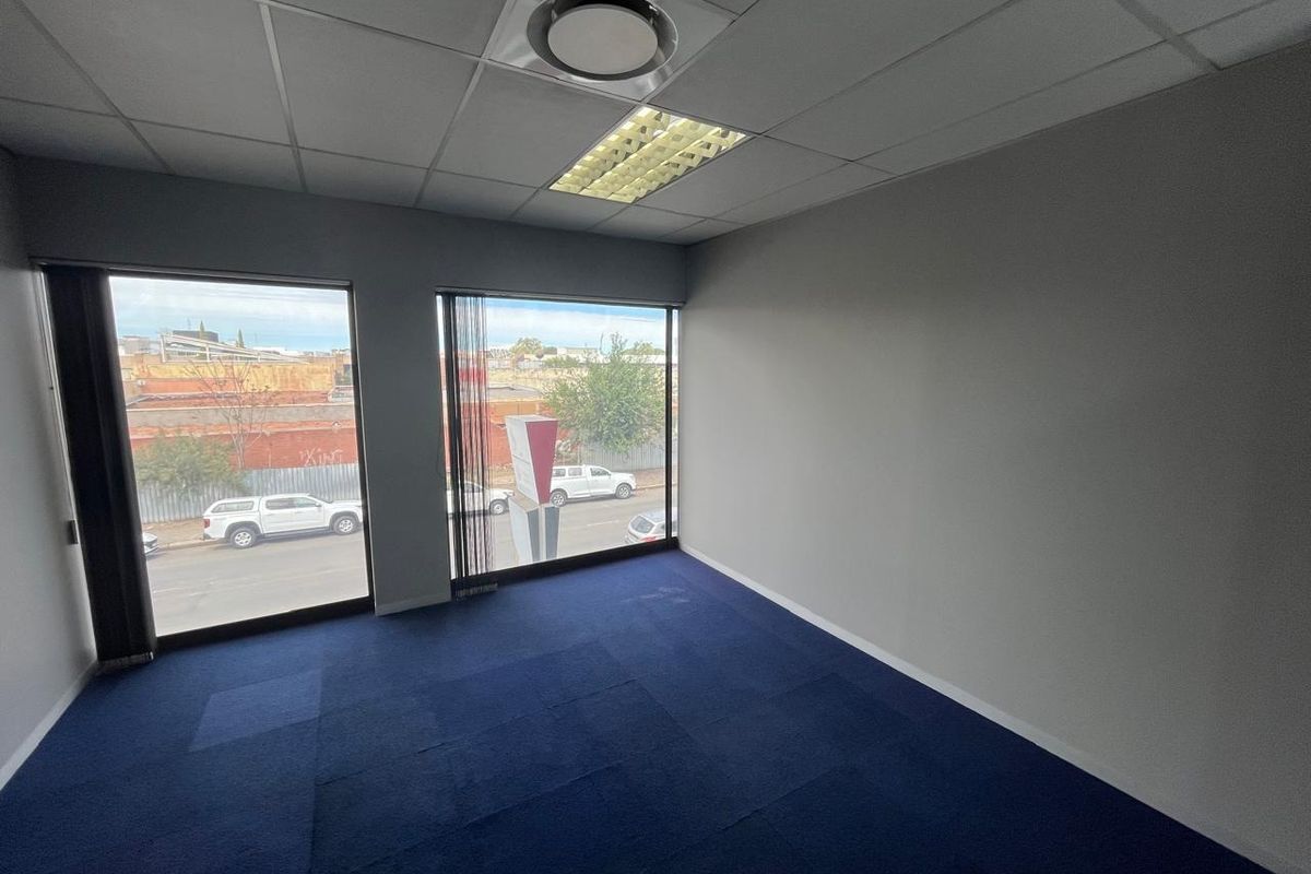 A vacant office space featuring blue carpet tiles, a suspended ceiling with recessed lighting, and large windows providing natural light and street views.