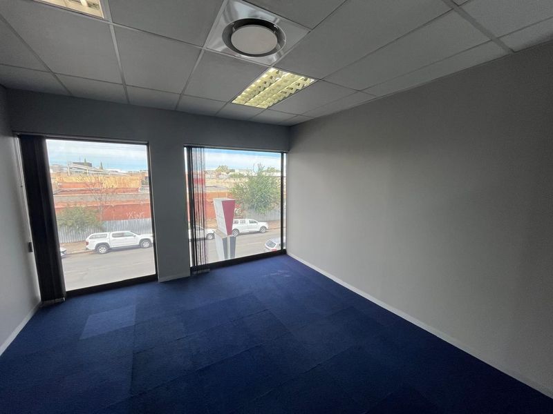 A vacant office space featuring blue carpet tiles, a suspended ceiling with recessed lighting, and large windows providing natural light and street views.