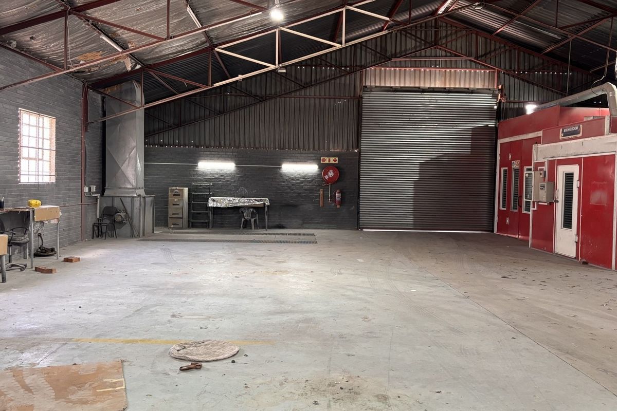 Spacious industrial workshop featuring a large roller shutter door, concrete flooring, exposed steel roof trusses, and an integrated spray booth facility.