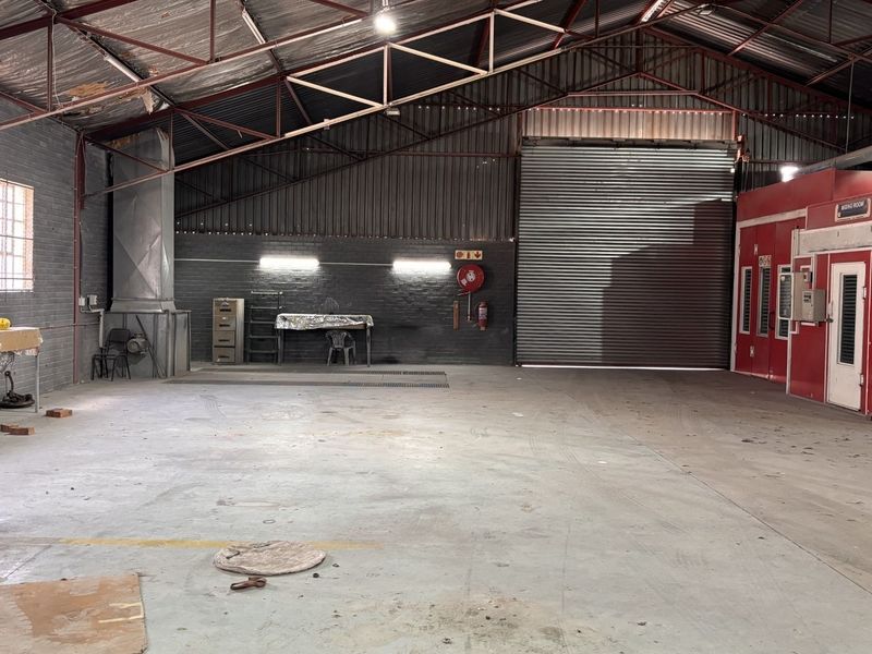Spacious industrial workshop featuring a large roller shutter door, concrete flooring, exposed steel roof trusses, and an integrated spray booth facility.