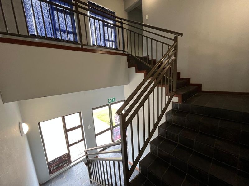 A bright, open-plan stairwell featuring dark tiled steps, a metal balustrade, and large windows providing natural light to the building entrance area.