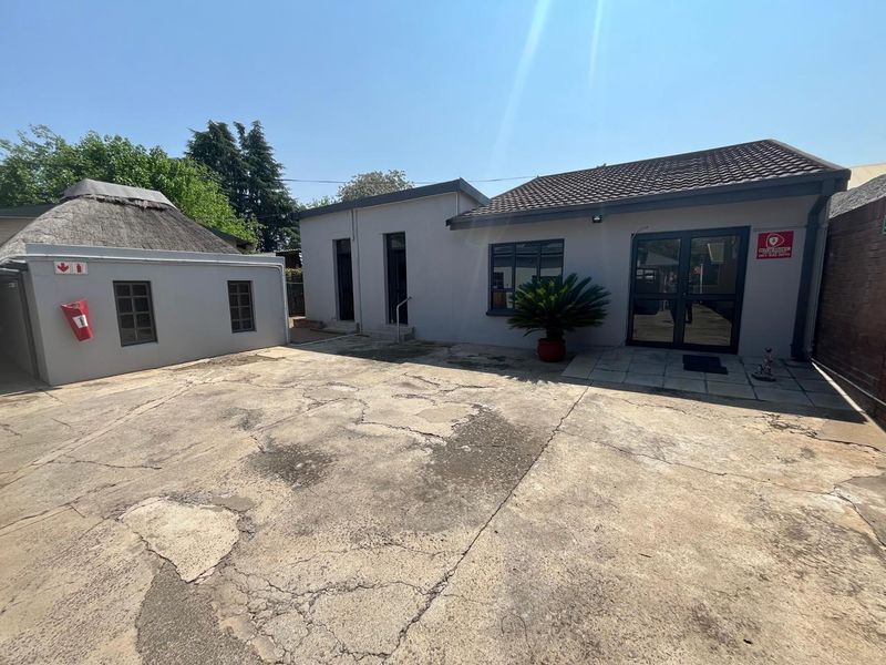Rear exterior view of the office property featuring a large concrete parking area, tiled roof sections, and a secondary thatched structure.