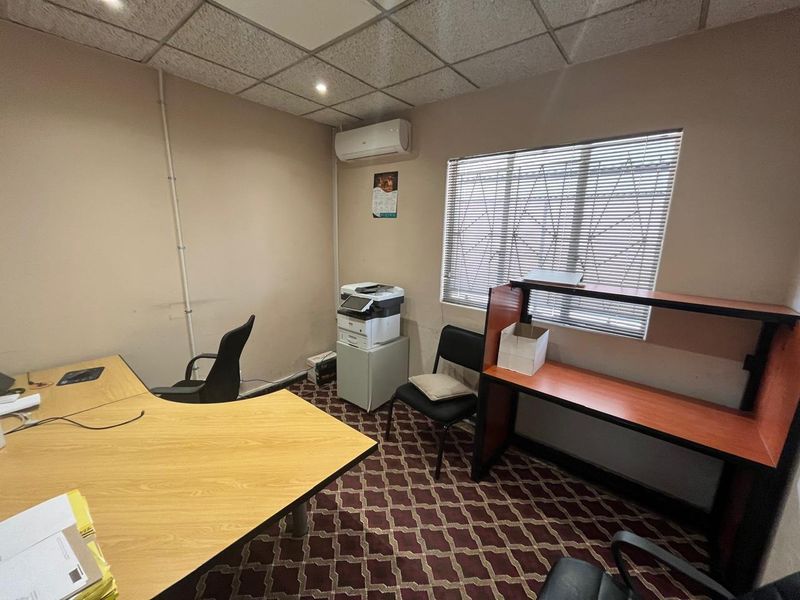 A functional office space featuring a suspended ceiling, recessed lighting, a wall-mounted air conditioning unit, and a window with security bars and blinds.