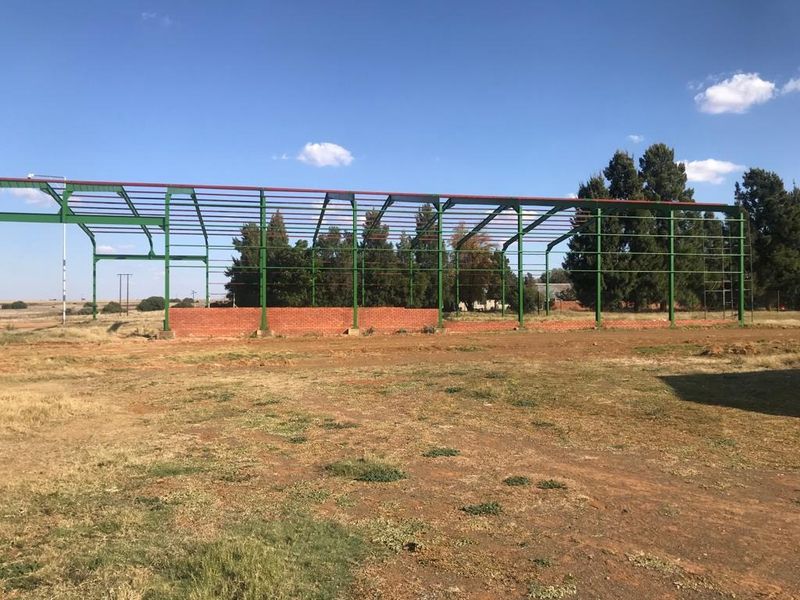 An incomplete steel portal frame structure with a low brick foundation wall, situated on an open farm plot.