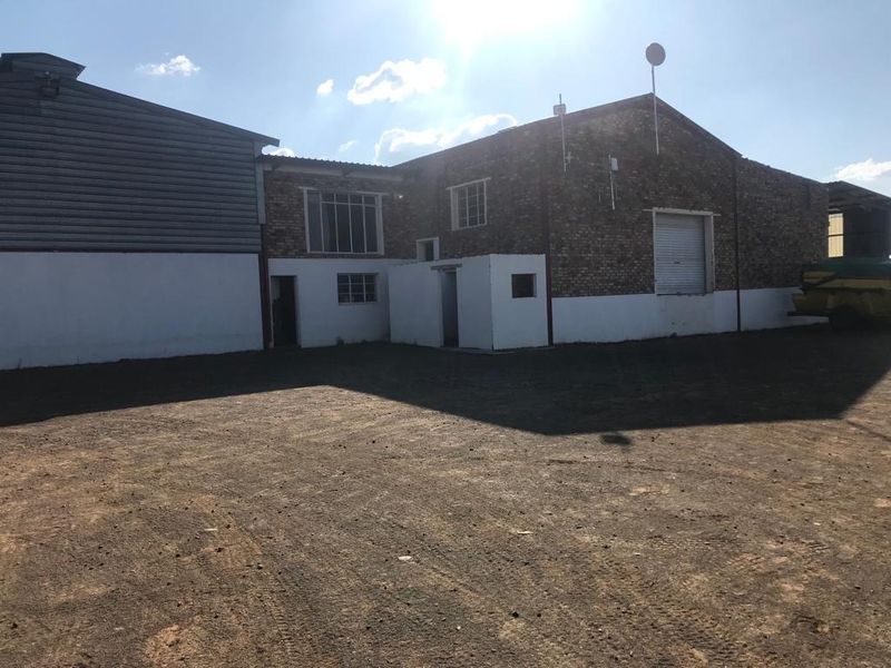 Rear view of a farm building featuring brick construction, metal roller shutter doors, and steel-framed windows set against an unpaved yard.