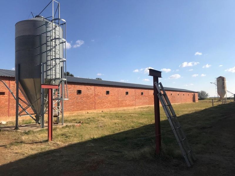 Exterior view of a farm property featuring a large brick barn, metal grain silo, and steel structural supports on the grounds.