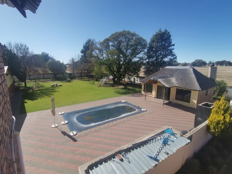 Aerial view of a small holding featuring a swimming pool, paved patio area, and a separate thatched-roof building set within a spacious garden.